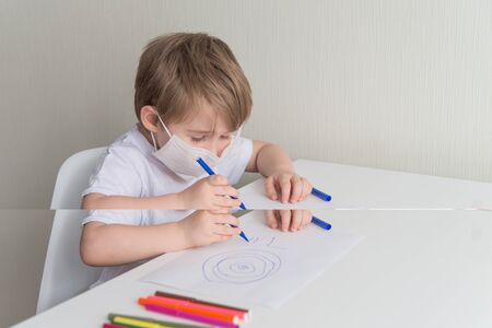Little boy in medical mask sits on the chair near the table and drawing with colour pensの写真素材