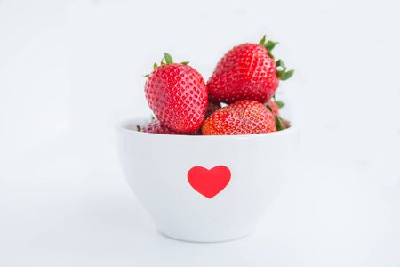 Fresh strawberries in white bowl with red heart on the white background close upの写真素材