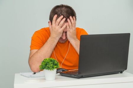 Tired overworked man in orange shirt holds his head sitting near laptop. Online work. Online education conceptの写真素材