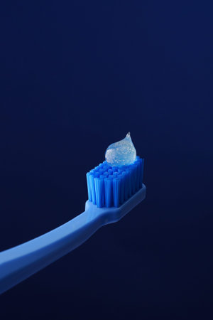 Blue plastic toothbrush on a dark blue background with a drop of transparent toothpaste. Oral...