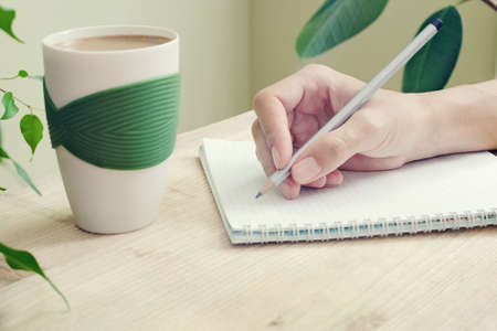 The hand of a woman with a pencil is written in a diary with spirals. Beside to the table is a cup of coffee and plant with green leaves. Side viewの写真素材