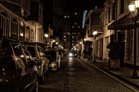 Street of the old night city with paving stone, lit by street lamps with parked cars.の写真素材