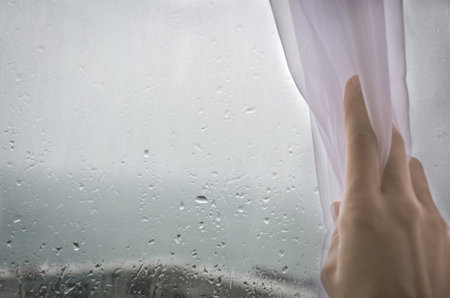 Woman's hand pulls the curtain off the wet rainy window. Empty copy space for text.