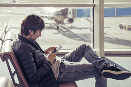 Young caucasian man in jeans and outerwear is sitting in chair airport waiting hall and using smartphone. Online remote banking, hotel booking, mobility, reading news, use of 3 G and 4 G internet networks. Traveling people. Airplane with jetway at background.の写真素材