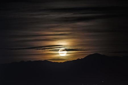 Night scene - full moon shining brightly in the sky with clouds on mountains background.