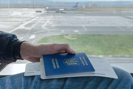 Manâs hand is holding Ukrainian foreign passport with tickets to airplane in background parked plane. Concept of  trip abroad, visa free regime, travel on vacation and freedom in traveling.の写真素材
