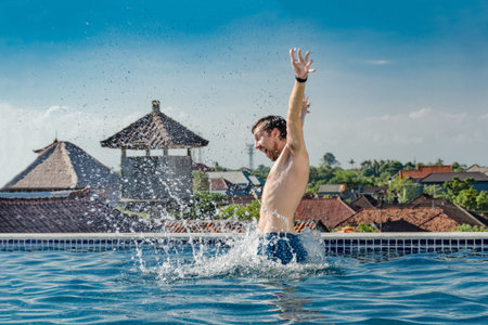 Summertime life - very happy, slender young smiling man, jumping out of pool water with splashes, on...