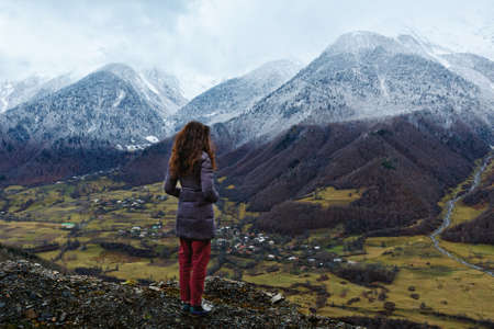 Young woman stands over a cliff and looks at snow-capped mountains, village in lower mount. Winter is coming, first snowfall, change of seasons. Georgia.の写真素材