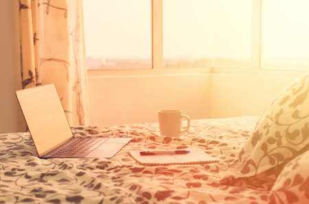 Sunny morning in modern apartment -  open laptop on bed opposite window, next to cup of coffee and notebook. Beginning of the day. Concept of working at home, productive day, planning. Photo toned in warm colorsの写真素材