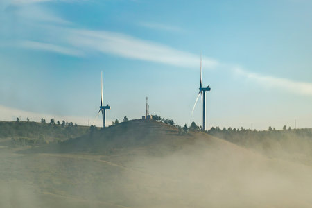 Wind farm of two wind turbine generators on top a hill, early morning, cloud of fog, meadow on...