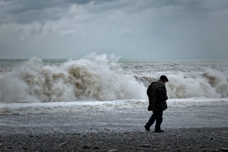 Batumi, Georgia, 2017-10-31: Dramatic, stormy weather, sea beach with big waves, single, lonely old man walking along coast, back view. Concept of loneliness, soul-searching, regret, loss of feeling in life.のeditorial素材