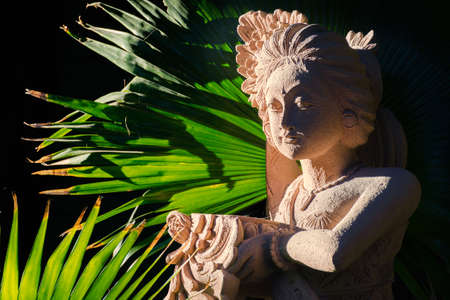 Traditional Balinese sandstone statue of woman,  welcome offering in her hands, illuminated by the sun among the palm trees.の写真素材