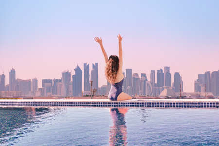 Woman with hands up enjoying morning view from roof top infinity pool at asia. Asian travel and vacation. Concept people at hotels with beautiful urban view.の写真素材