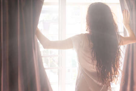 Sunny morning - a woman opens the curtains and the sun shines through the window.  Back view. Blurred photoの写真素材