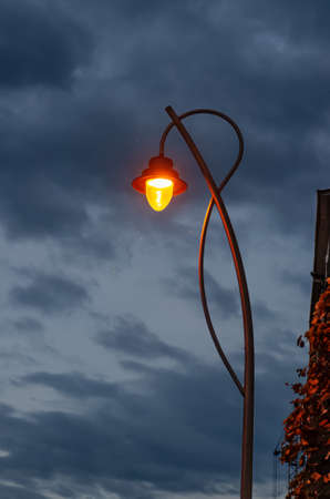 Street lantern shines with warm light against evening cloudy sky background. Midges fly in the light glowの写真素材