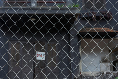 Blurred building with dark walls behind an iron mesh fence.  Sale sign