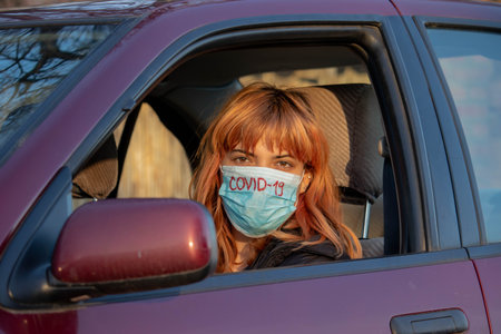 Girl wearing medical mask in the car. Woman wearing mask, written COVID-19, to protect from corona virus. Driving car with mask during virus pandemic. Mask must be put on in everyday activities.の写真素材