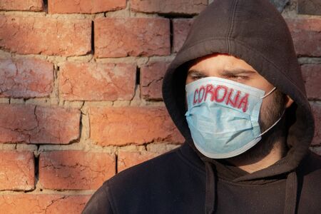 Man in hood  with mask to protect him from Coronavirus. Corona virus pandemic. Corona written on mask. Young man with mask standing next to brick wall. Person in hood with medical mask.の写真素材