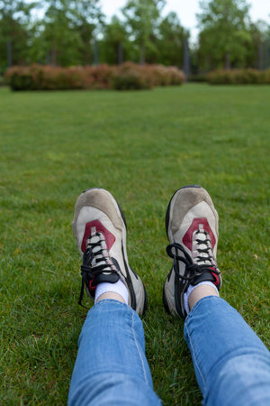photo of women's legs sitting in jeans and stylish leather sneakers on the grass, in the park, on a...