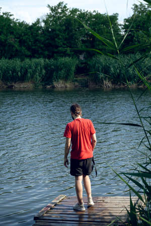 A man in a red T-shirt and shorts stands on a wooden pier and looks into the distance.の写真素材