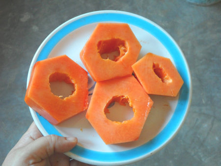 Four papaya slices cut in the shape of nuts, and served on a white plate with blue rim, over a grey background; you can see the thumb of the human hand holding itの写真素材