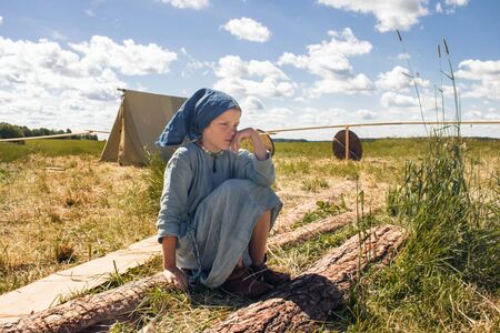 STARAYA LADOGA, RUSSIA -06.23.2019: Campground of participants of the Festival of Historical Reconstruction. A girl in a medieval folk costume sits on a log in front of a bonfireのeditorial素材