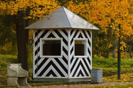 Black and white striped guardhouse on the background of yellow trees and a white marble bench in the park in autumnの写真素材