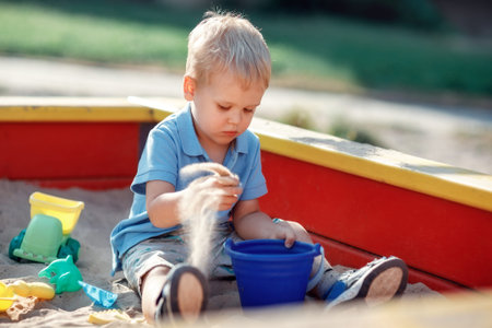 Toddler boy is playing in a red and yellow edge sandbox with sand toys. Focused baby, wearing blue...