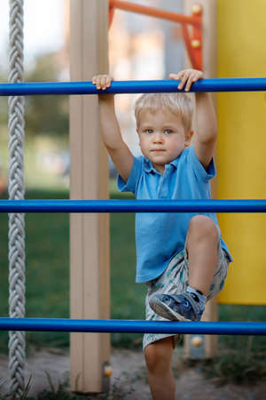 Small boy is climbing on a blue playground ladder. Looking at camera. Active child. Modern playgroundの写真素材
