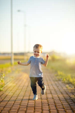 Cheerful little boy with striped shirt runs down the sidewalk. Sunny summer evening, backlighting.の写真素材