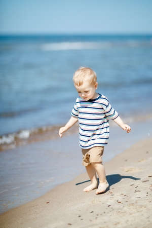Barefoot little boy walks on the seaside at noon near the water. Child on vacation in summer at the sea on good weather.の写真素材