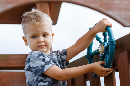 Beautiful child is driving a ship wheel in a playground. Kid wearing camouflage feels relaxed in a...