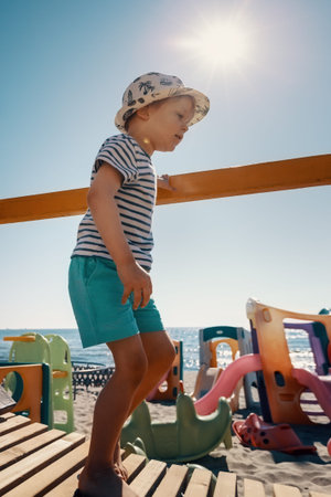 A cute boy in a hat walks on a wooden bridge to the beach. A playground, sea and sun rays in the backgroundの写真素材