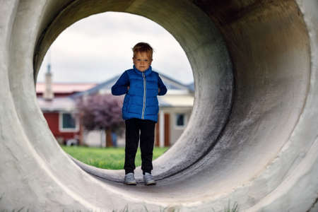 A cute kid boy, wearing a blue vest, stands in a large concrete tunnel. Children's games in the yard.の写真素材