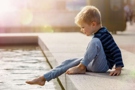 A cute little boy, barefoot, wants to climb into the water at the city fountain during the summer.の写真素材