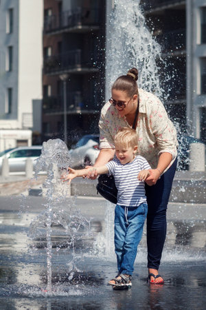 Mom and her son, in the city center, by a fountain of water. The child is afraid to touch the water so that it does not get wet, the mother encourages it.の写真素材