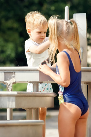 A happy boy and a cute girl in a blue bathing suit play with a water tap in a city park. Vertical.の写真素材