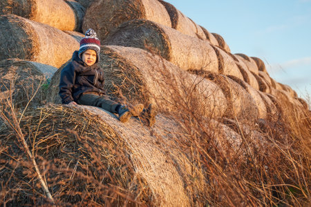 A cute little boy in autumn clothes and a knitted hat poses against a background of golden hay bales. Horizontal photo.の写真素材