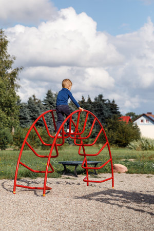 A little very brave boy climbs a metal, big red arched ladder on the playground. The concept of parental care and safety in children's play.の写真素材
