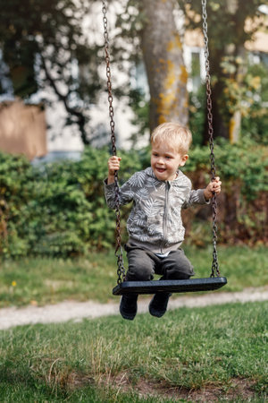 Little boy having fun on a swing on the playground in public park on sunny summer day. Happy child enjoy swinging. Active outdoors leisure for child in city.の写真素材
