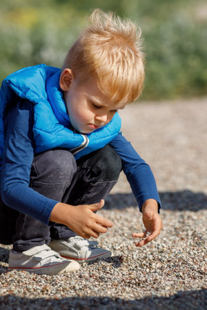 A little boy with a blue vest plays outdoors with small pebbles. The child explores the ground with his own hands, he is very interested in small pebbles.の写真素材