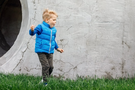 A cute boy wearing a blue vest plays outdoors on the grass near a cement wall with a big hole.の写真素材