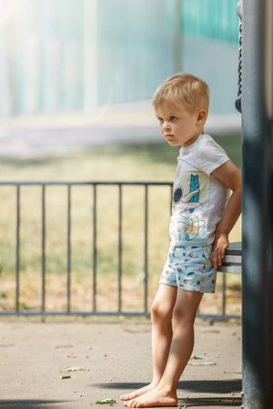 A blond, barefoot boy in a white shirt takes a break on an outdoor playground on a hot summer day.の写真素材