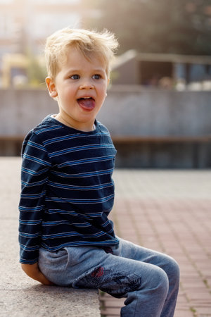 A portrait of a little naughty, sympathetic boy sitting by a city fountain, the child poses, showing his tongue.の写真素材