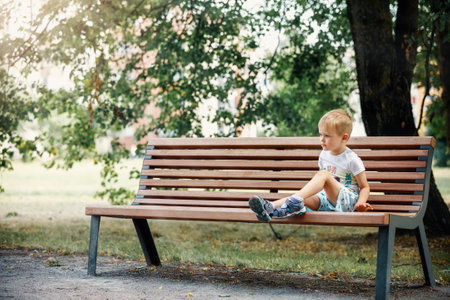 Little boy sitting on a bench. Baby alone in the city. Large tree and sunlight in the background.の写真素材