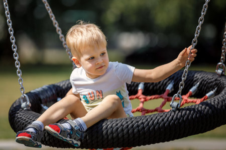 A cheerful little boy swings in the park on a swinging rope swing. Round black and red seat for children's swing.の写真素材