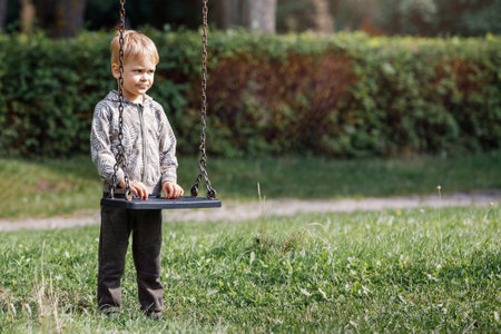 An attractive little boy stands next to a swing with chains in a city park, a child getting ready to swing. Horizontal photo.の写真素材