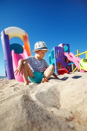 A little boy in a hat sits on a sand and play with it, beach and colourful playground in a background. A beautiful summer day, free time and a carefree childhoodの写真素材
