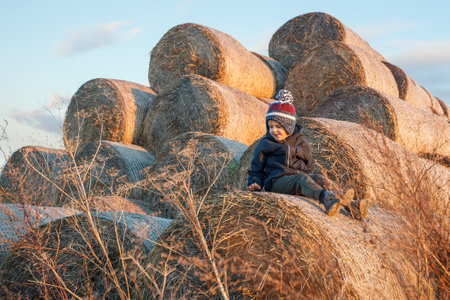 A child wearing warm autumn clothes and a hat sits in the evening light and plays on a pile of golden straw rolls in the village.の写真素材