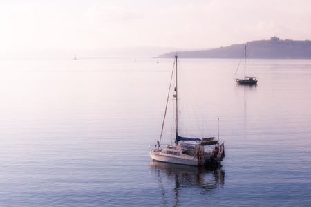 High-key image of yachts moored in calm sea at St Mawes in Cornwall as the morning mist lifts.の写真素材